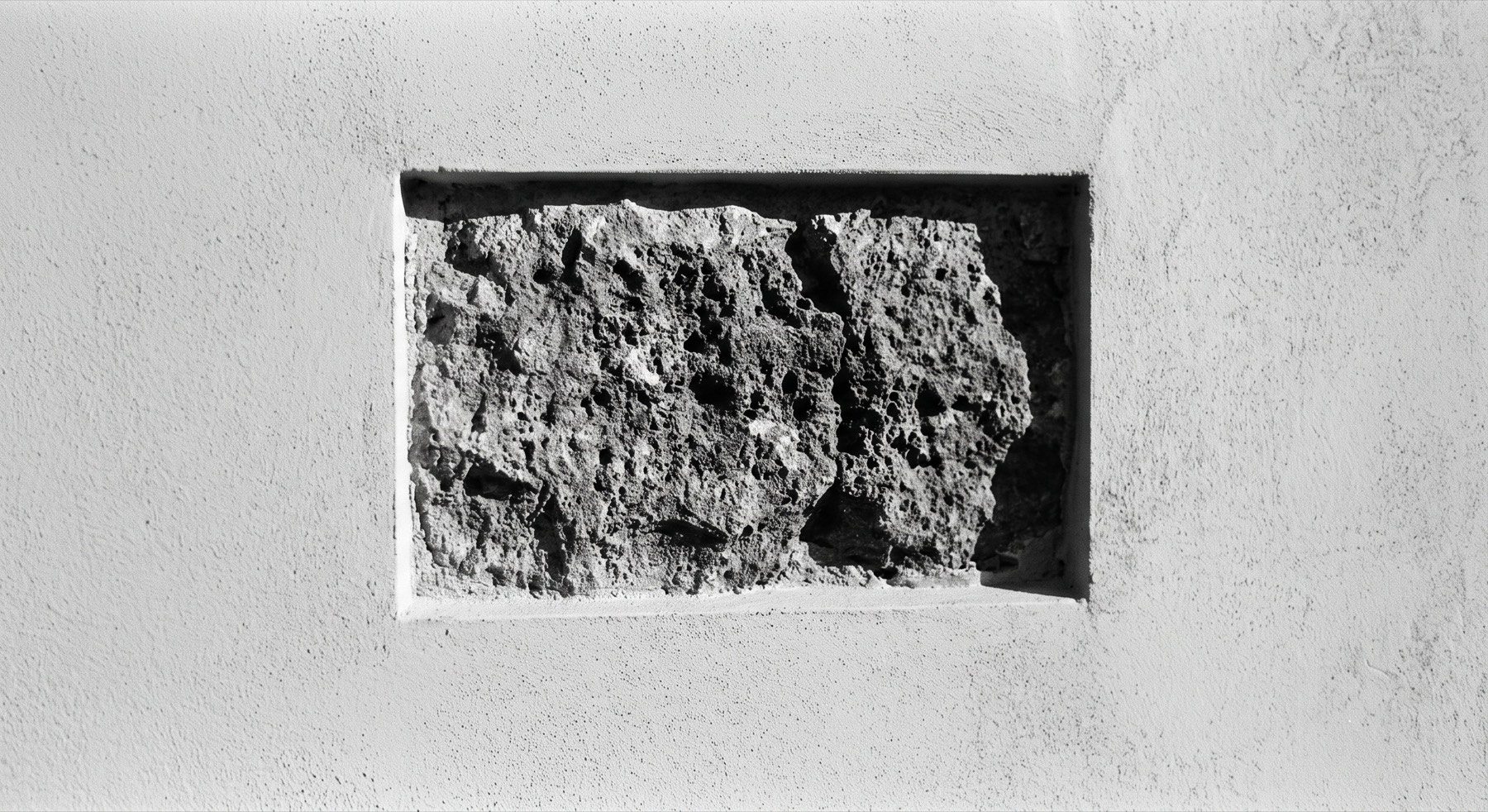 A smooth white rendered wall with a rectangular section cut away to leave one rough, weathered stone exposed in the middle