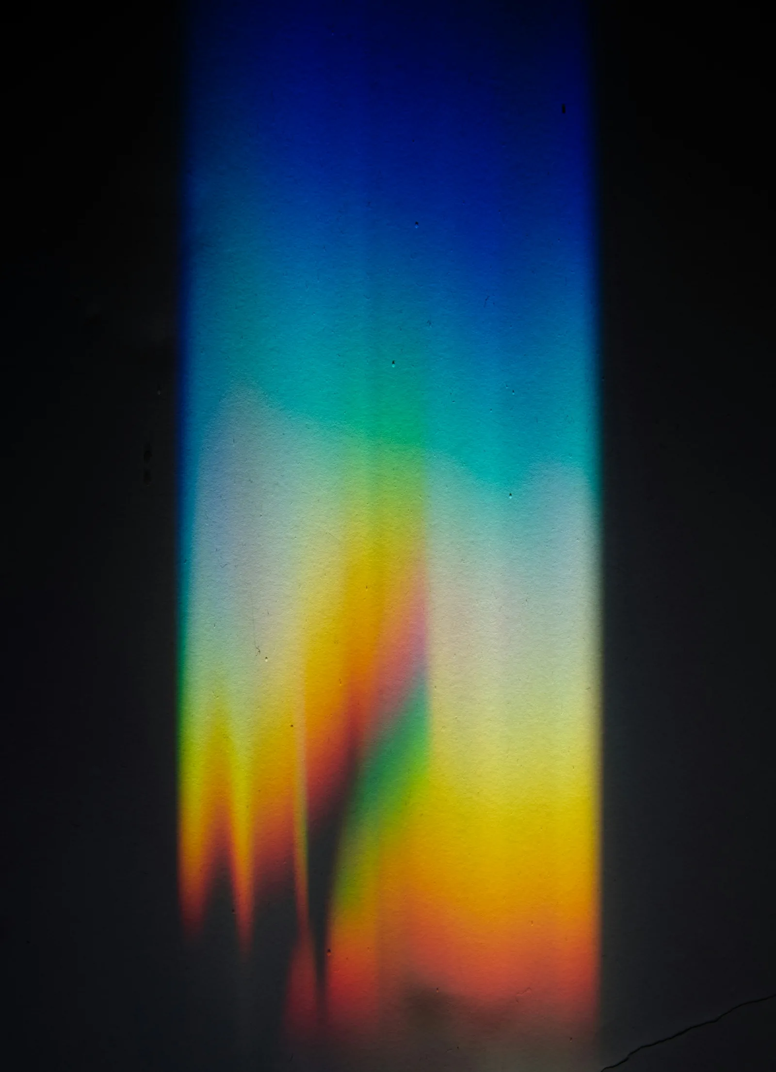 A band of refracted rainbow light falling across a dark wall, blue through to orange, New Beach journal
