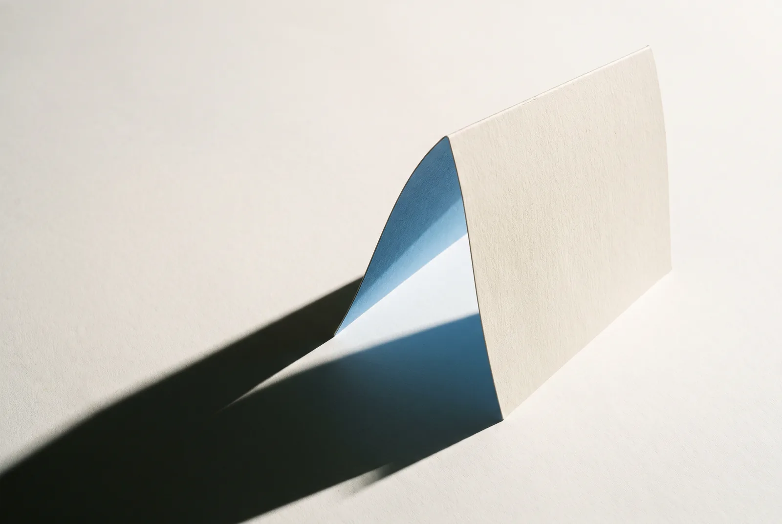 A curved sheet of cream card standing on a pale surface, blue gradient on its inner face, New Beach journal