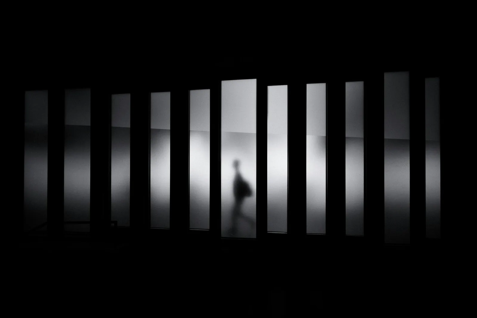 A blurred figure walking behind tall backlit frosted panels in a dark room, New Beach journal