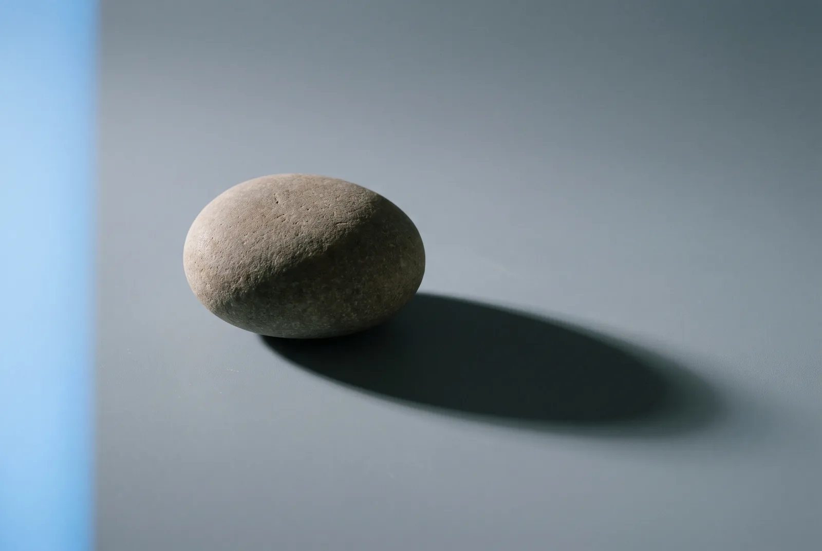 A single smooth pale pebble on a grey surface, casting a long shadow beside a strip of blue light, New Beach journal