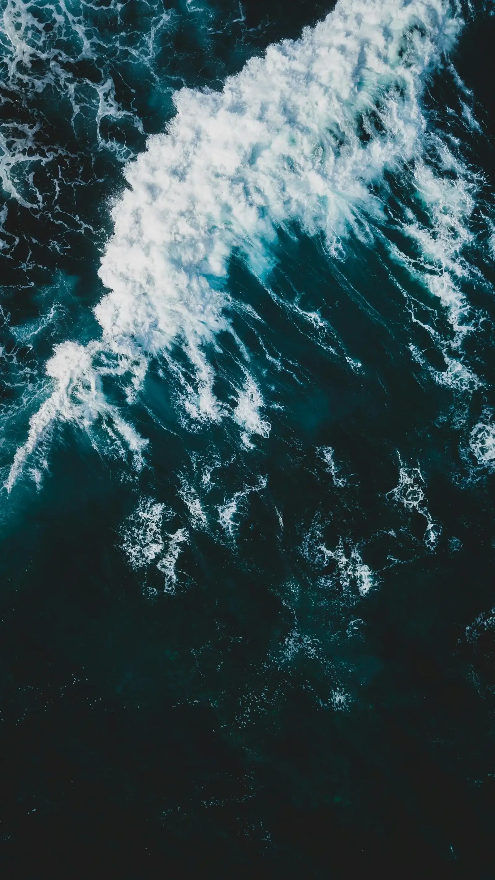 Aerial view of a wave breaking, white foam spreading across dark teal water, New Beach journal