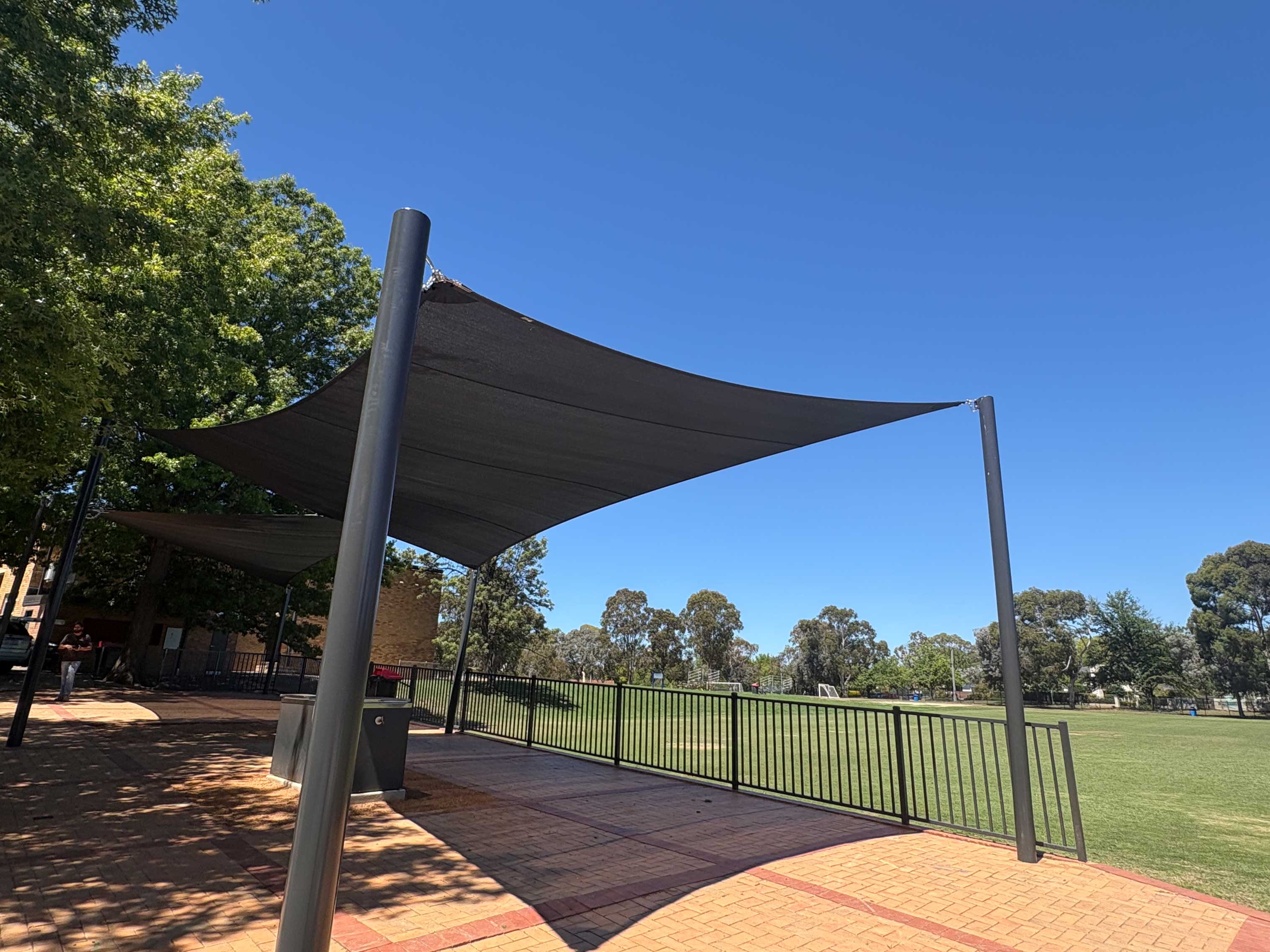 Large shade sail on steel posts over a paved outdoor area beside a school oval