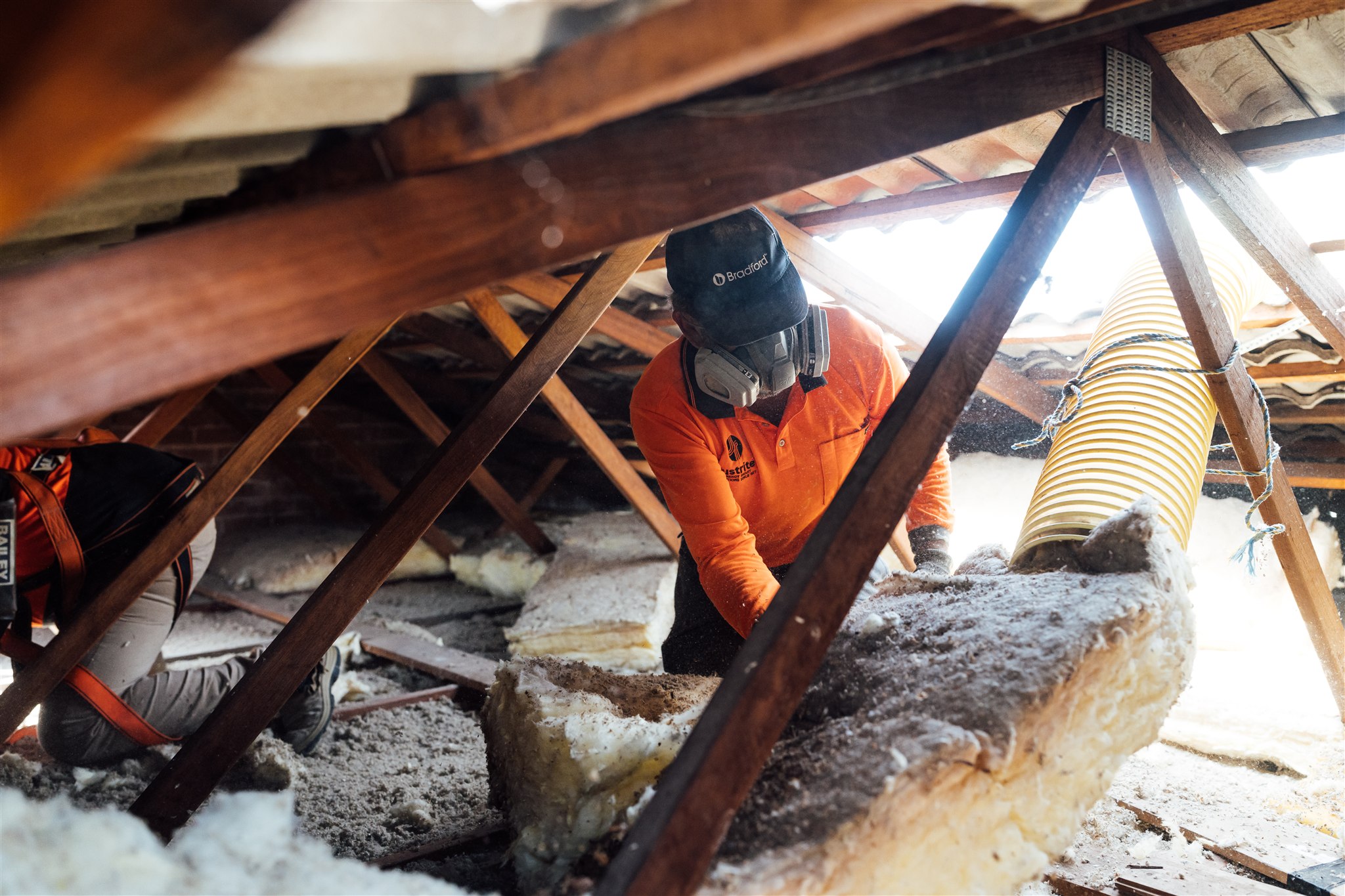 Installer in orange hi-vis laying insulation batts in a roof cavity between timber trusses