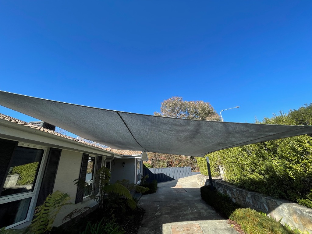 Charcoal shade sail stretched over a driveway beside a home under a clear blue sky