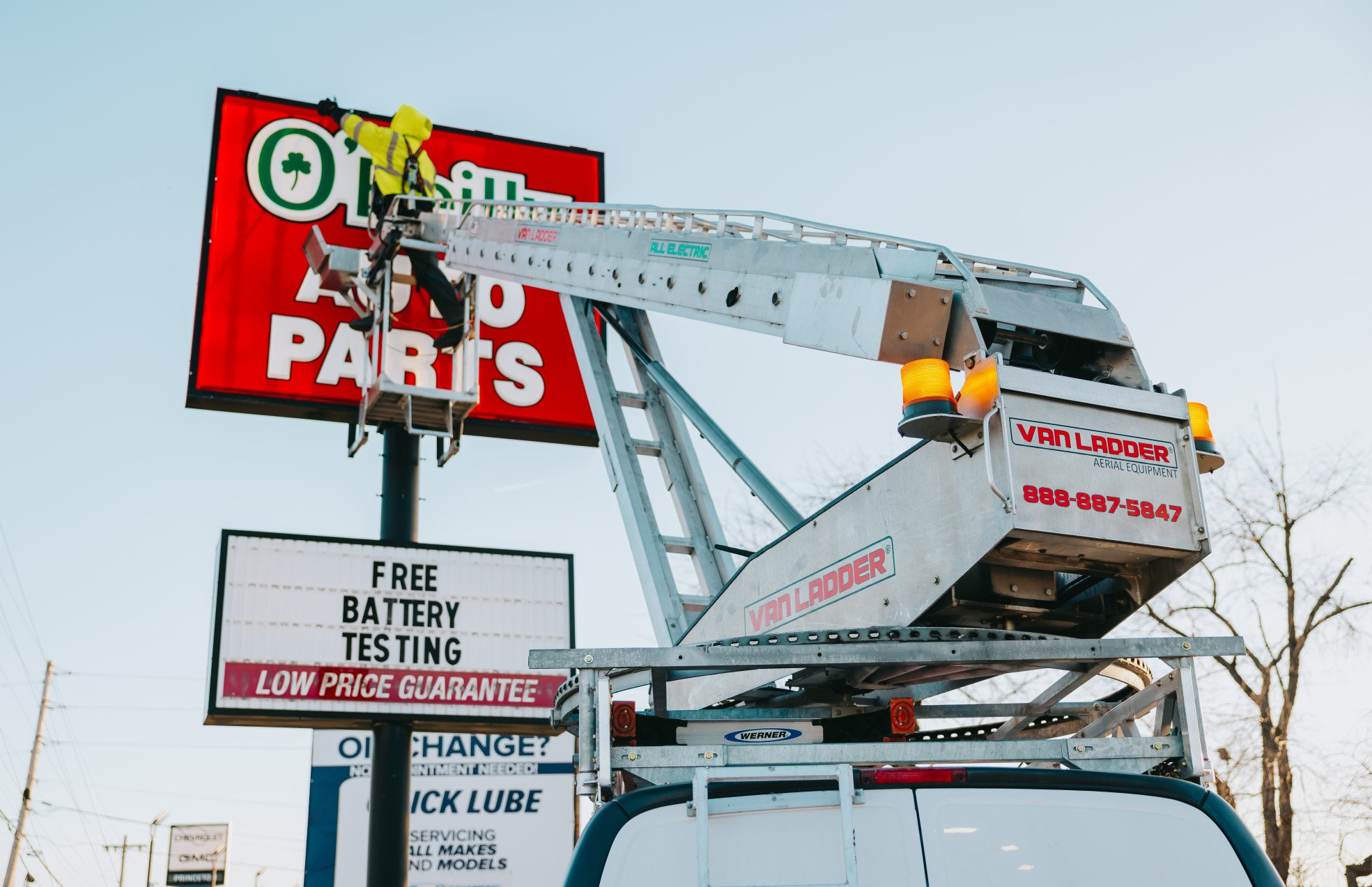 Rick Signs Van Ladder bucket truck approaching a tall O’Reilly Auto Parts pole sign with FREE BATTERY TESTING signage in Chicago