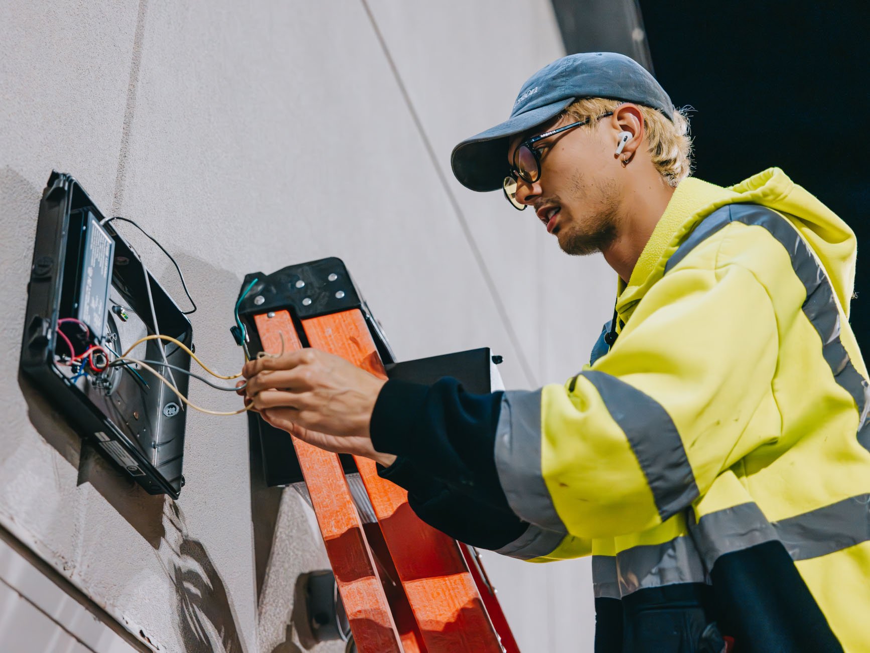 Worker in yellow jacket fixing wires in an open electrical box on a wall using a ladder.