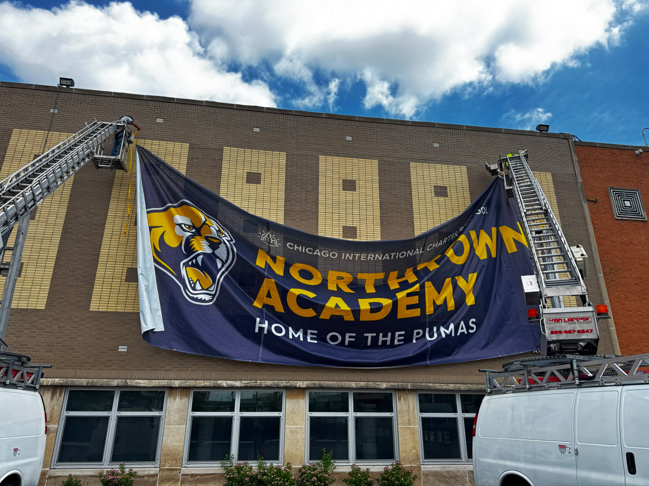 Workers hang a large Northtown Academy banner on a brick building using ladder trucks.