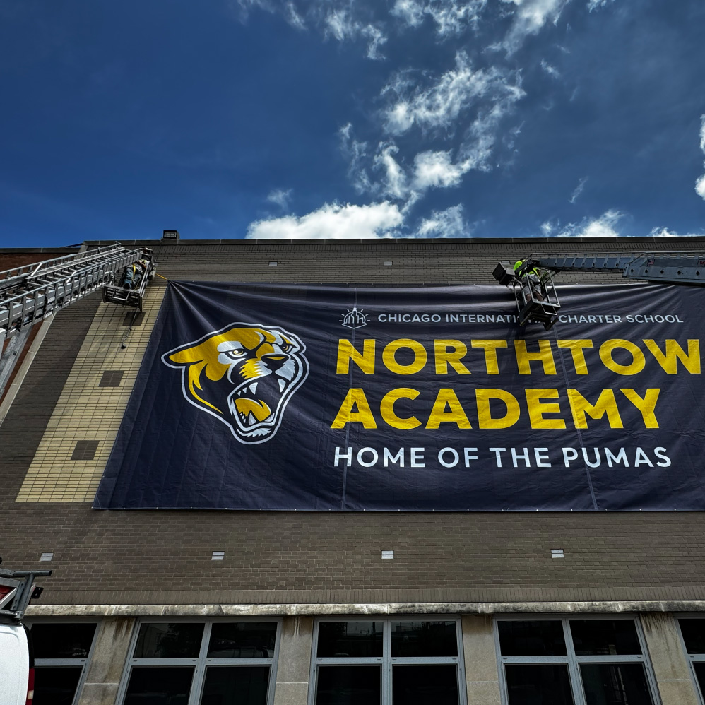 Workers install a large Northtown Academy banner with a puma logo on a brick building wall.
