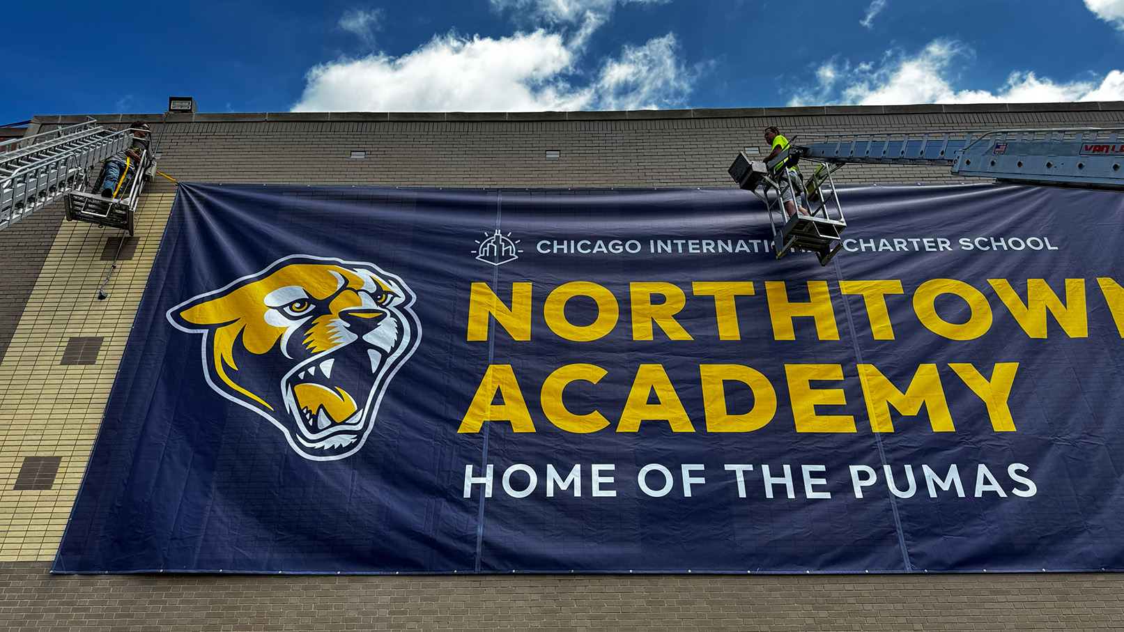 Workers installing a large banner for Northtown Academy with a puma logo on a brick wall.