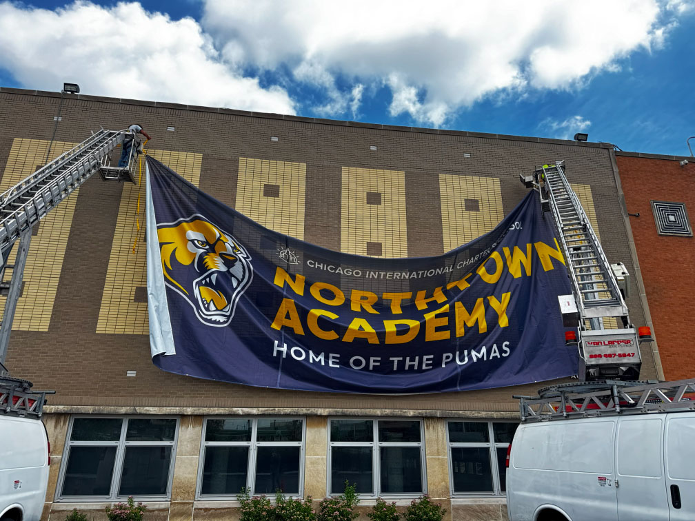 Two workers hang a large Northtown Academy banner showing a puma mascot on a brown brick building.