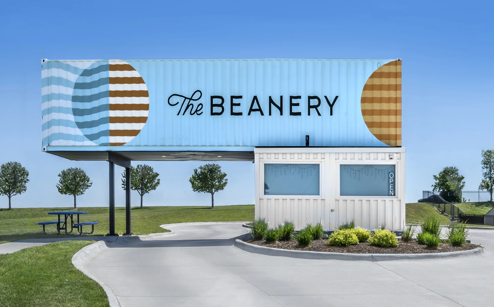 A light blue shipping container coffee shop named The Beanery with a white booth and open sign.