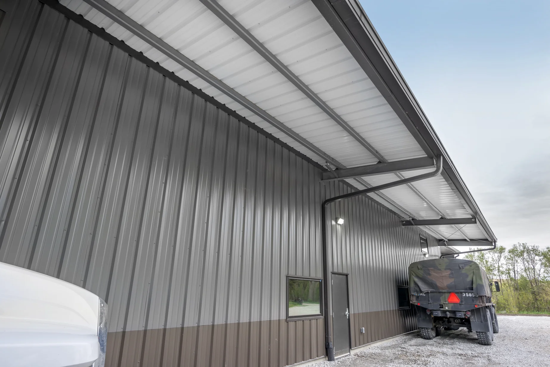 Exterior detail of a steel building with a canopy eave, metal siding, and parked vehicles nearby.