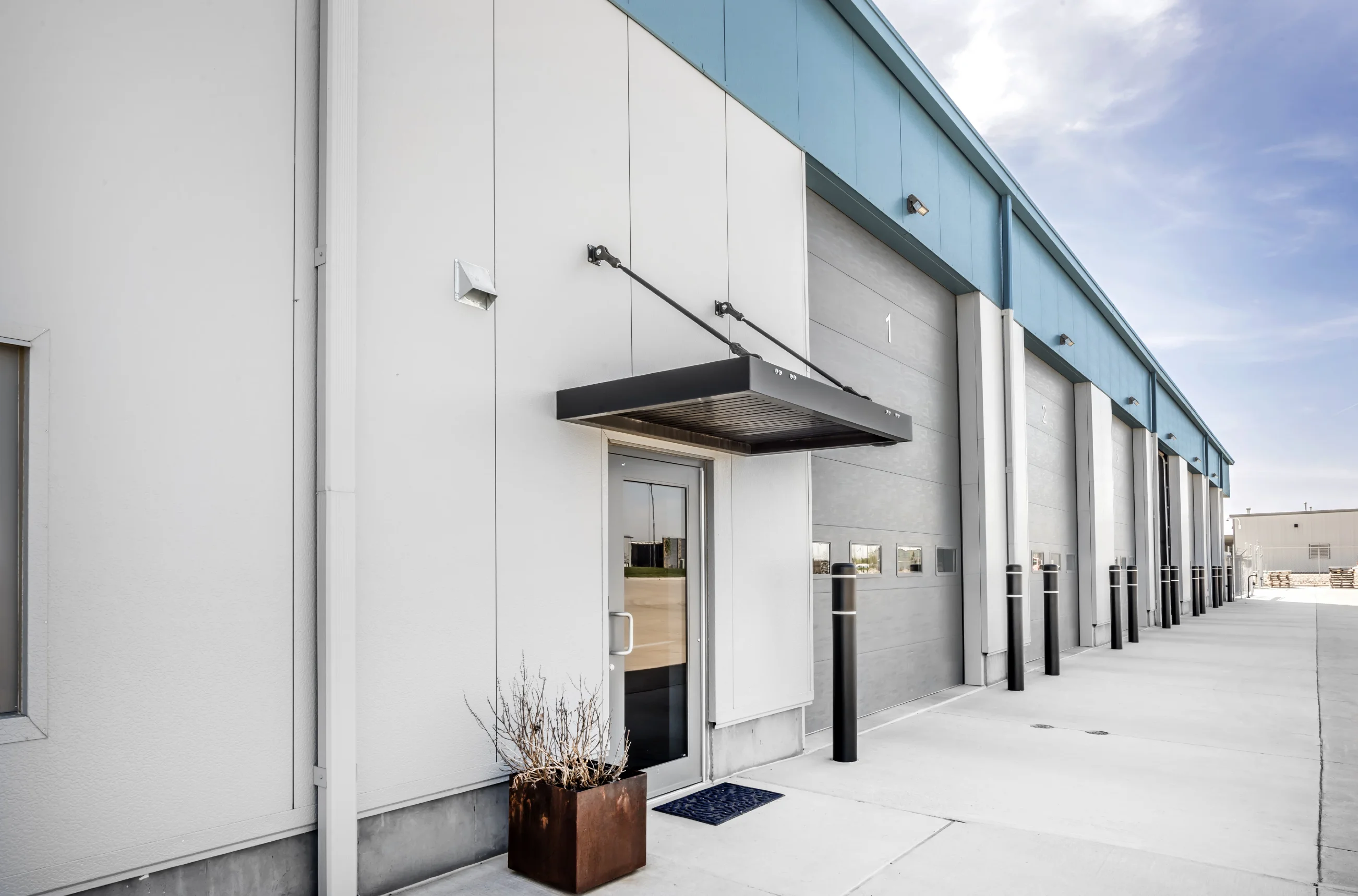 Commercial building entrance with a glass door, black awning, planter, and loading dock doors numbered 1 and 2.