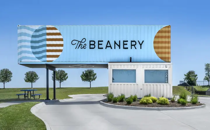 A light blue shipping container coffee shop named The Beanery with a white booth and open sign.