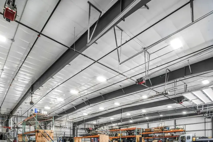 Interior of a large warehouse with metal beams, storage racks, equipment, and bright ceiling lights.