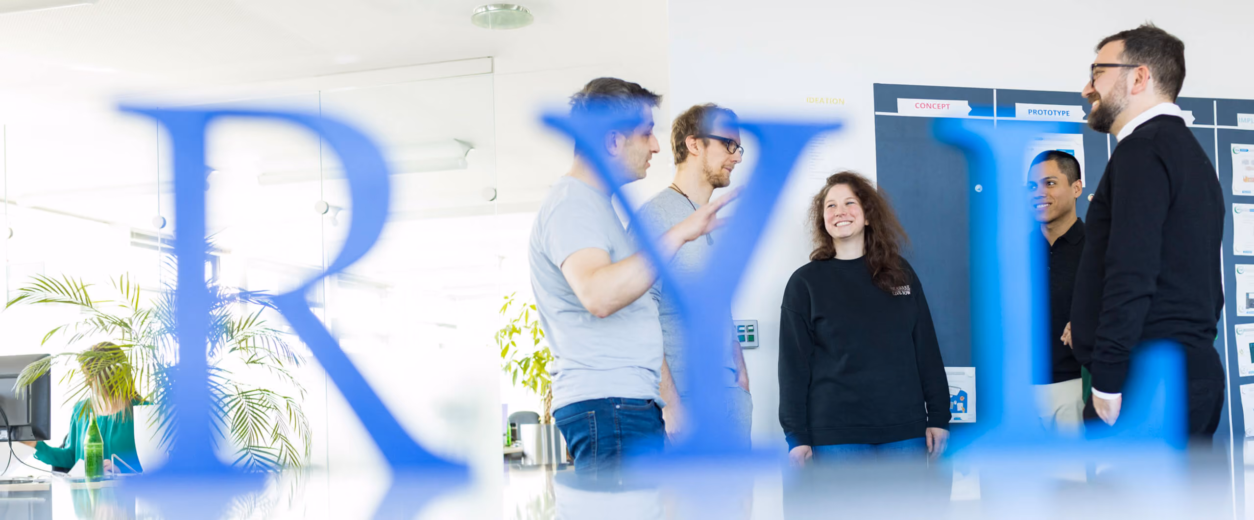 Fünf Personen führen ein Gespräch in einem modernen Büro hinter großen blauen Buchstaben.