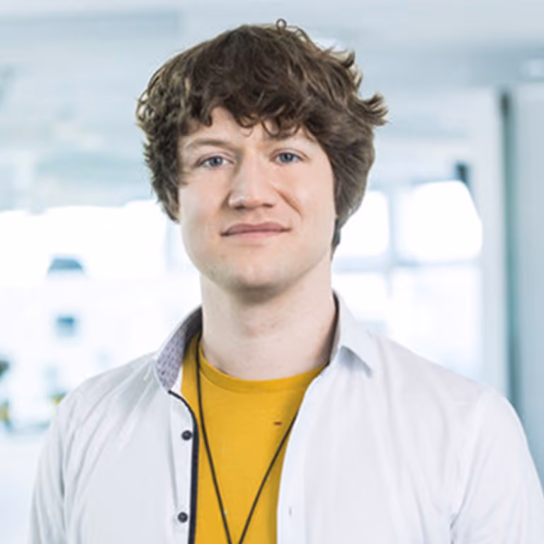 Junger Mann mit lockigem Haar trägt weißes Hemd und gelbes T-Shirt in hellem Raum.