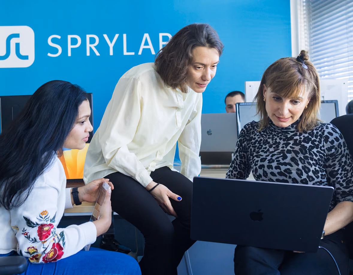 Drei Frauen sehen gemeinsam auf einen Laptop in einem Büro mit blauem Sprylar-Logo.