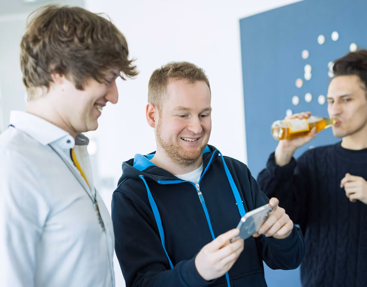 Drei Männer, zwei schauen lächelnd auf ein Smartphone, der dritte trinkt aus einer Flasche.