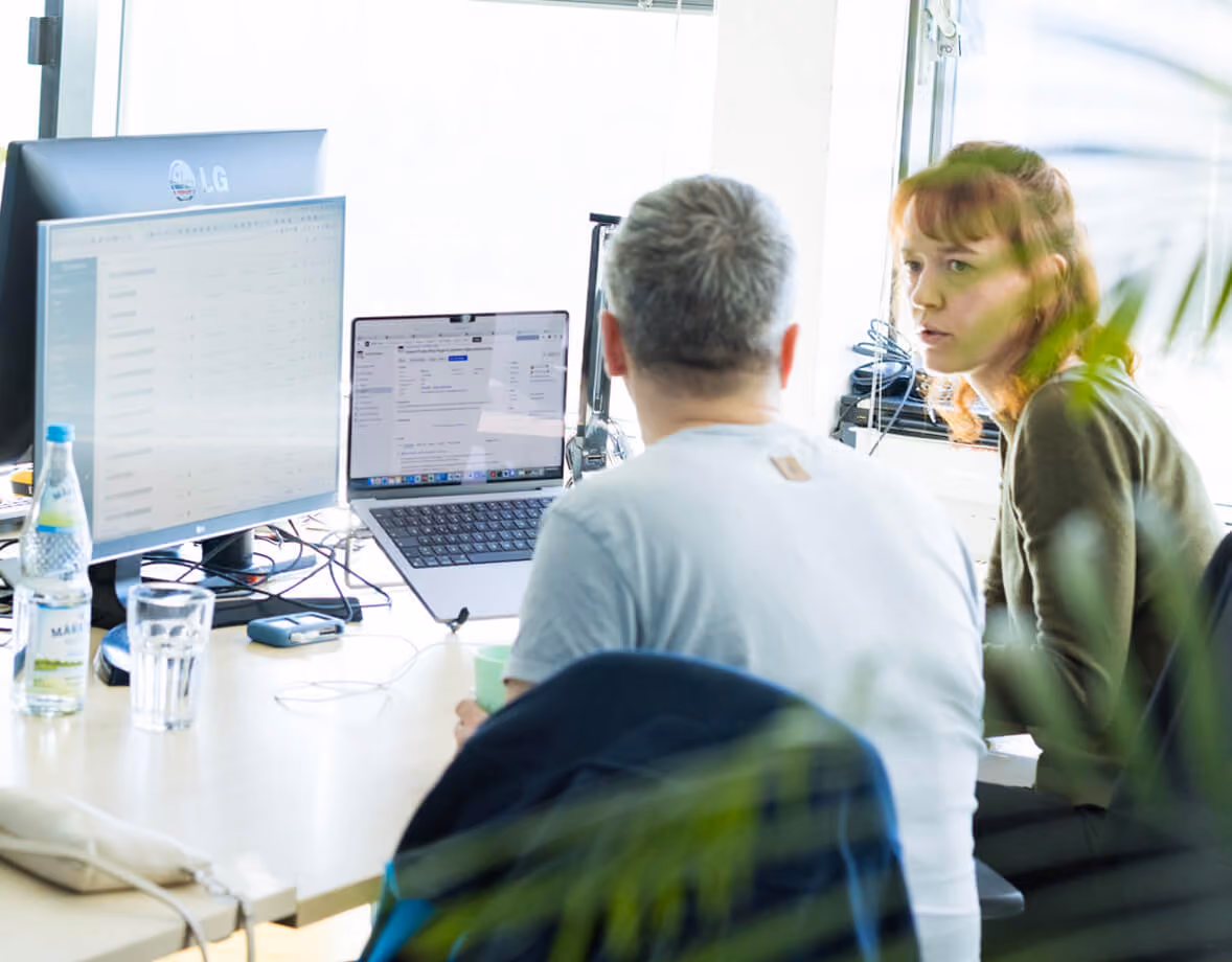 Zwei Personen besprechen Arbeit vor mehreren Bildschirmen in einem hellen Büro.
