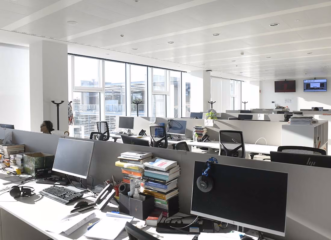 Helles Büro mit mehreren Schreibtischen, Computern, Büchern und einer Person am linken Rand.