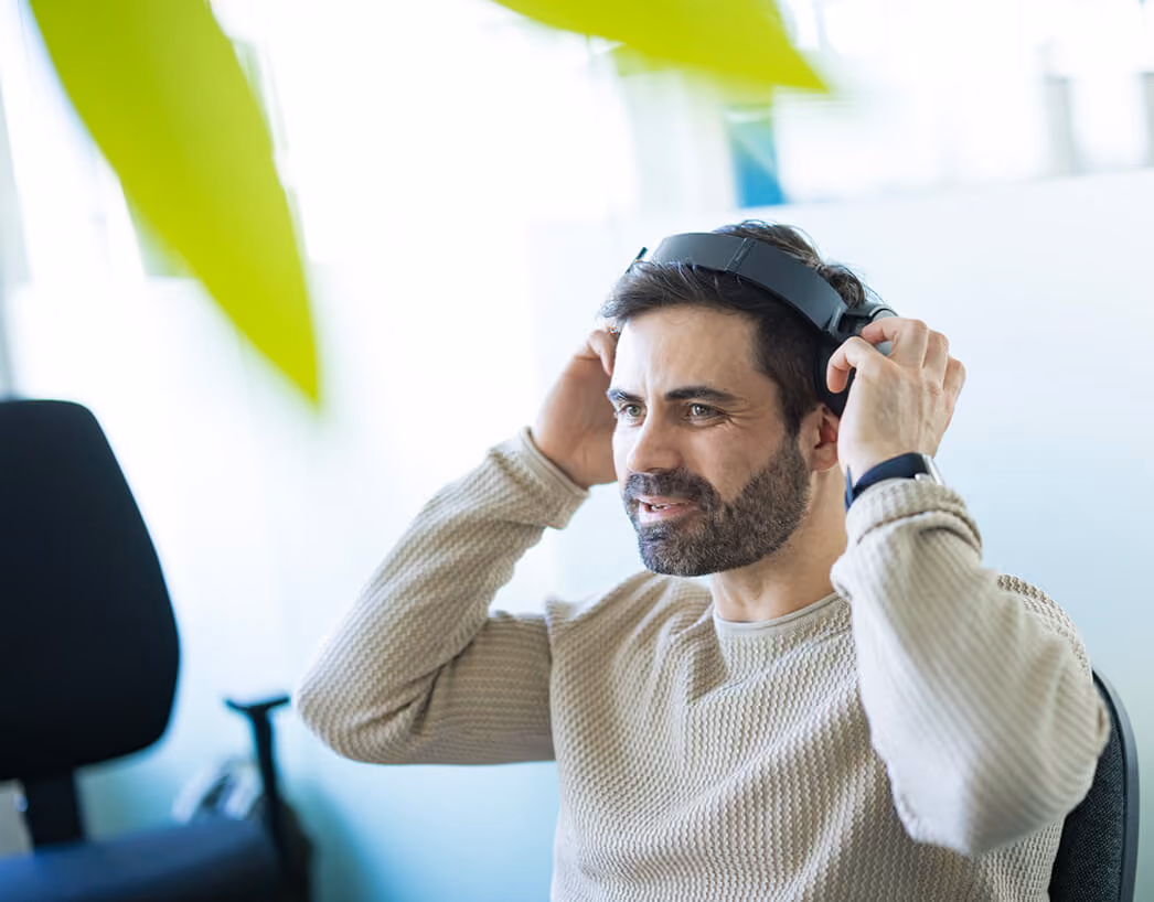 Mann mit dunklem Haar und Bart setzt Kopfhörer auf und sitzt in einem modernen Büro.