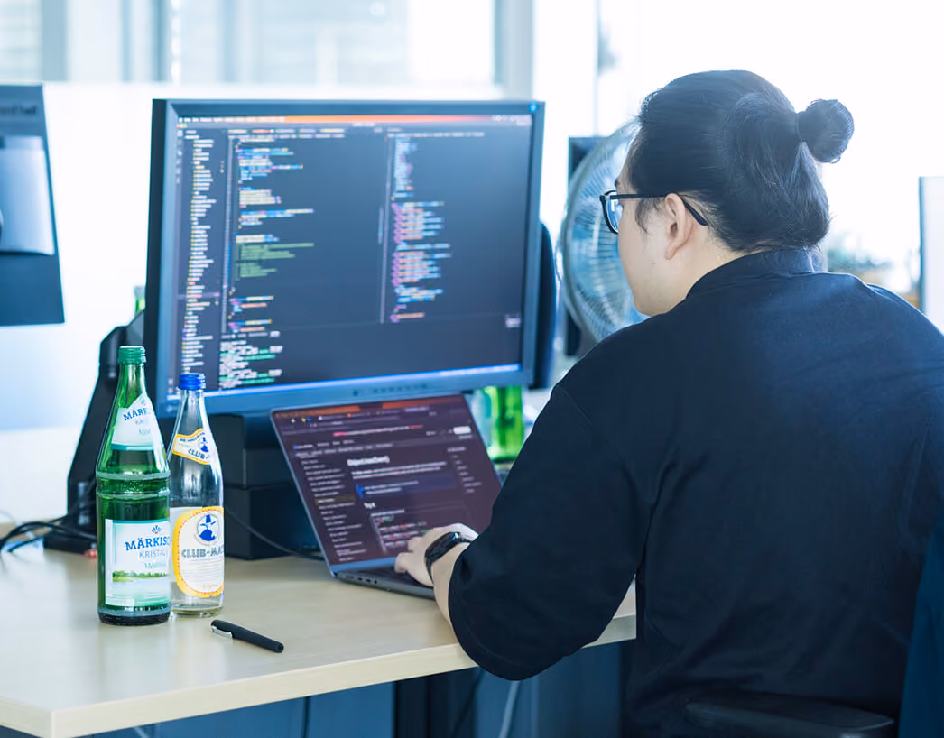 Person sitzt am Schreibtisch vor zwei Bildschirmen, auf einem ist Code sichtbar.
