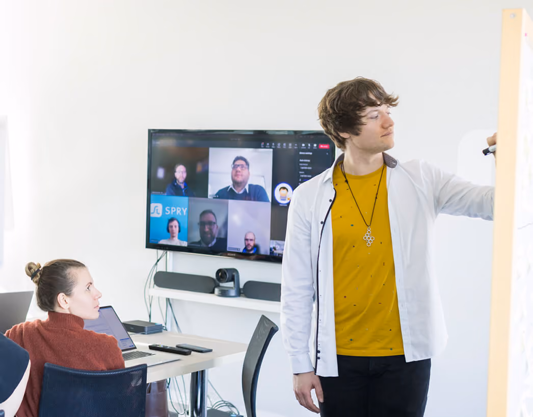Junger Mann schreibt auf Whiteboard, Frau sitzt am Tisch vor Laptop, Videoanruf mit mehreren Personen auf Bildschirm.