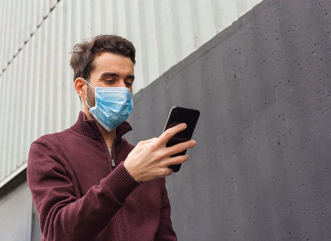 Mann mit Gesichtsmaske liest auf seinem Smartphone vor einer grauen Wand.