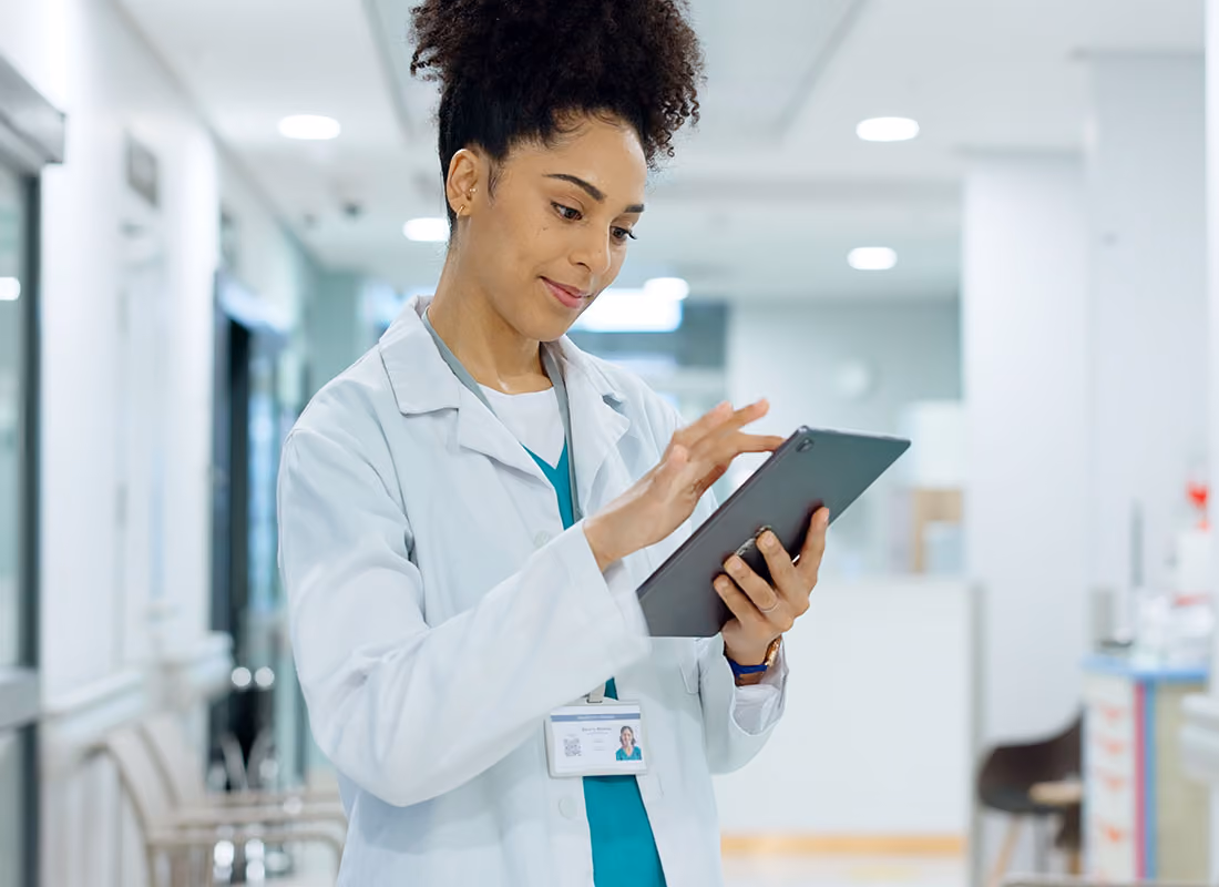 Ärztin mit Locken in weißem Kittel benutzt ein Tablet im hellen Krankenhausflur.