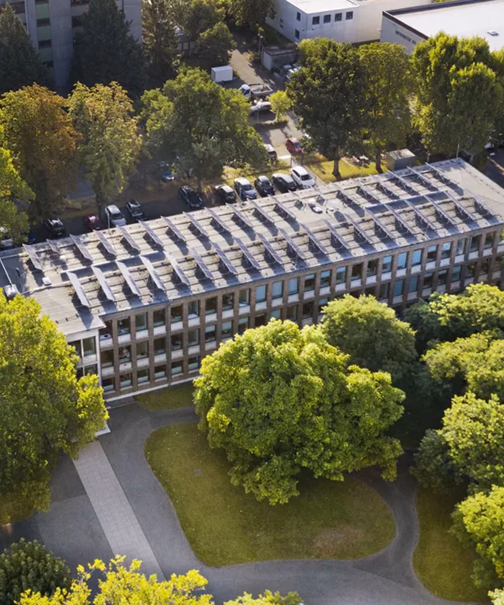 Luftaufnahme eines Gebäudes mit Solaranlagen auf dem Dach und umgeben von Bäumen und Parkwegen.