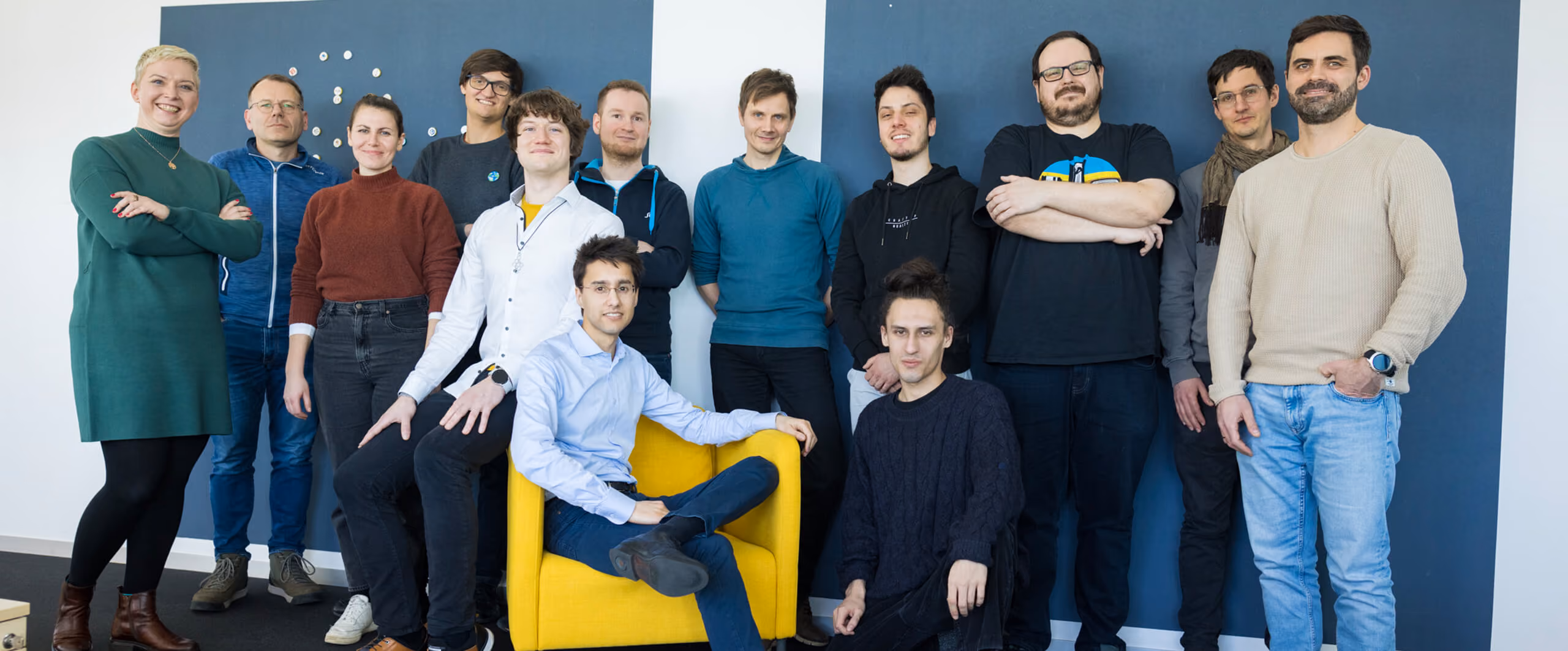 Gruppenfoto von 13 lächelnden Personen vor blau-weißem Hintergrund, einer sitzt in einem gelben Sessel.
