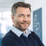 Porträt eines lächelnden Mannes mit kurzen braunen Haaren und Bart, der einen Pullover trägt.