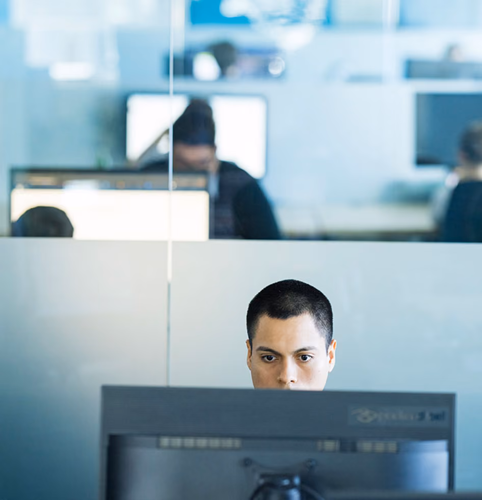 Person arbeitet konzentriert an Schreibtisch mit Computer in modernem Büro mit weiteren Personen im Hintergrund.