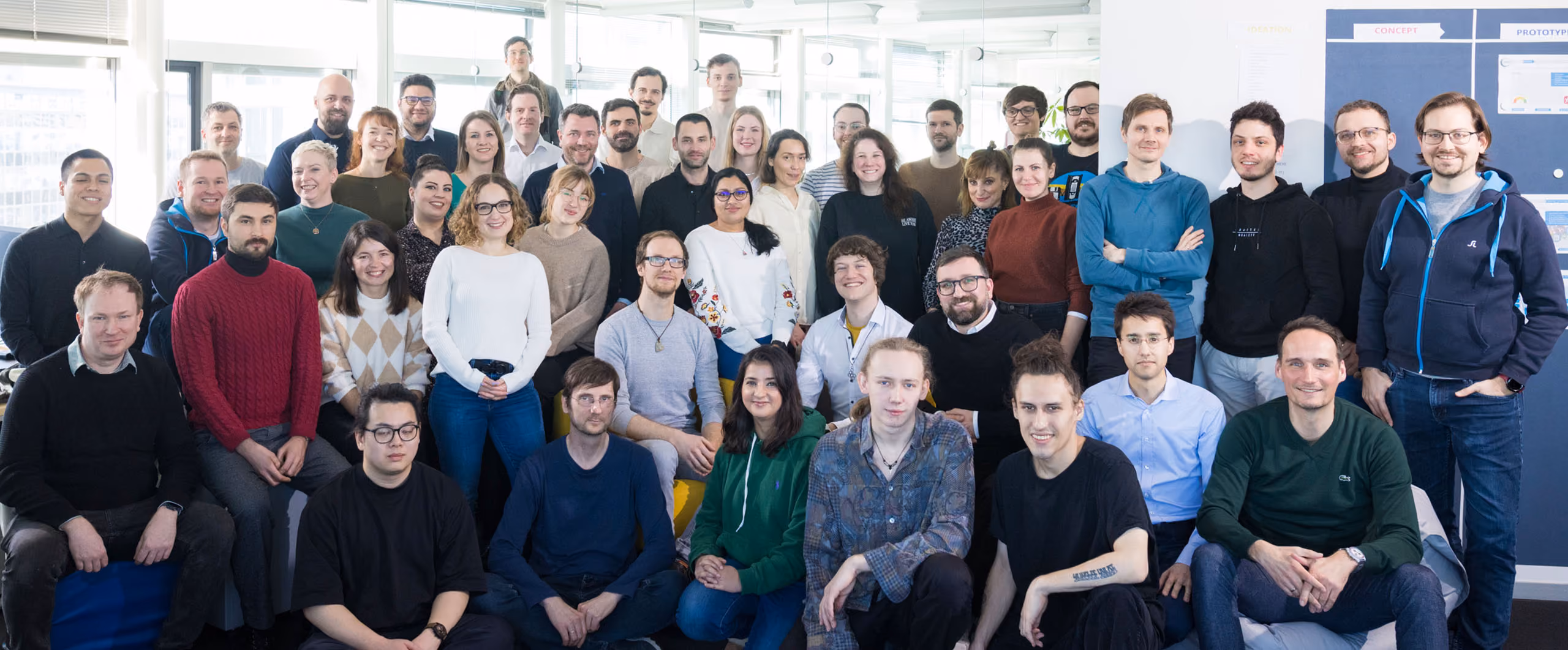 Gruppenfoto von 42 Mitarbeitern in einem hellen Büro mit Fenstern im Hintergrund.