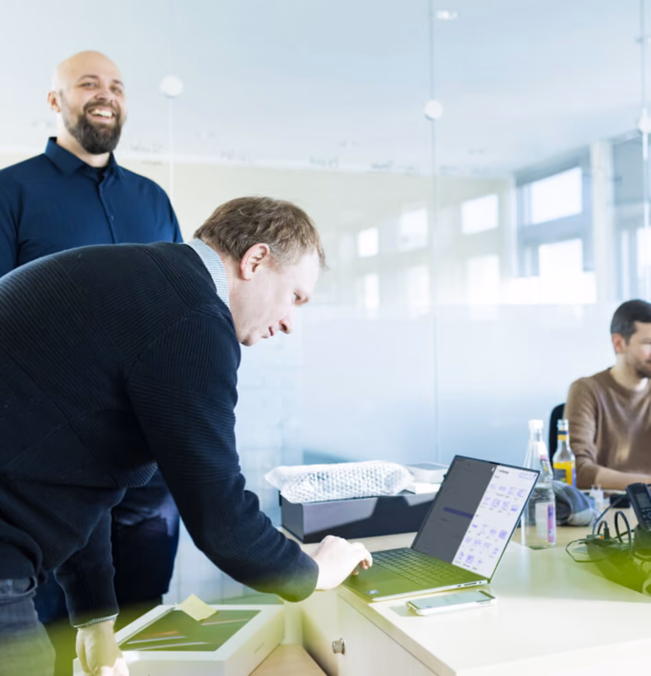 Zwei Männer am Schreibtisch, einer beugt sich zu einem Laptop, der andere lacht im modernen Büro.