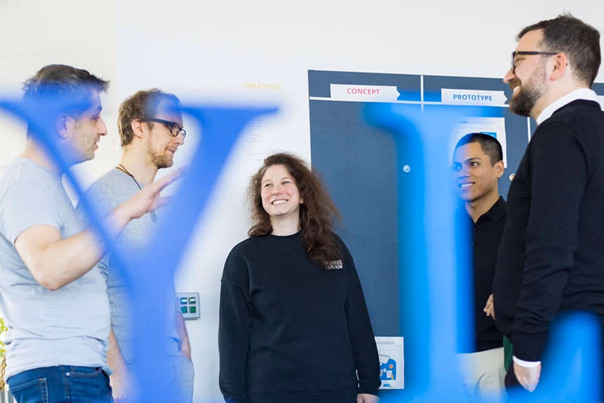 Fünf Personen stehen im Kreis und unterhalten sich vor einer Wand mit Beschriftungen Konzept und Prototyp.