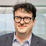 Mann mit kurzen lockigen dunklen Haaren, Brille, hellblauem Hemd und dunklem Sakko vor Fenster.