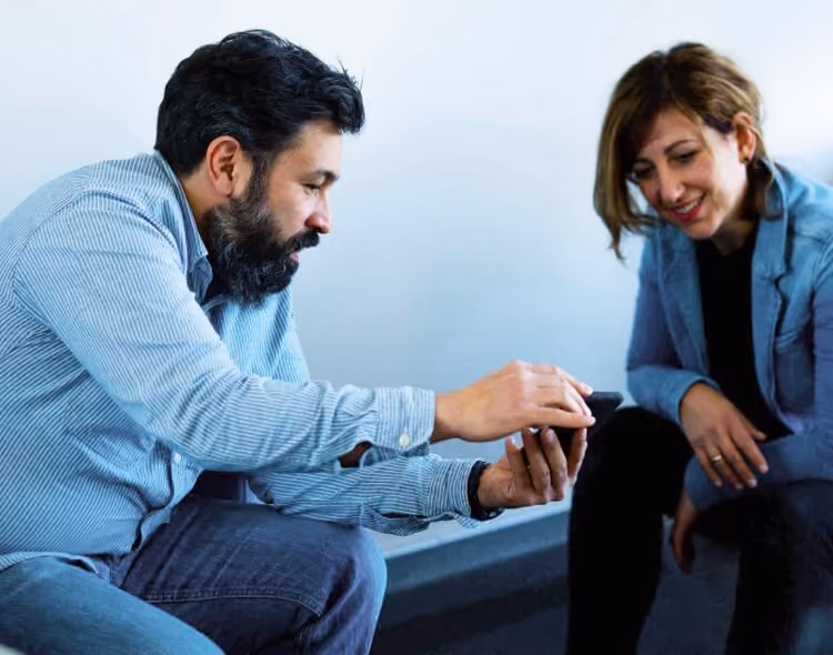 Ein Mann zeigt einer Frau auf einem Smartphone etwas, beide sitzen und lächeln.