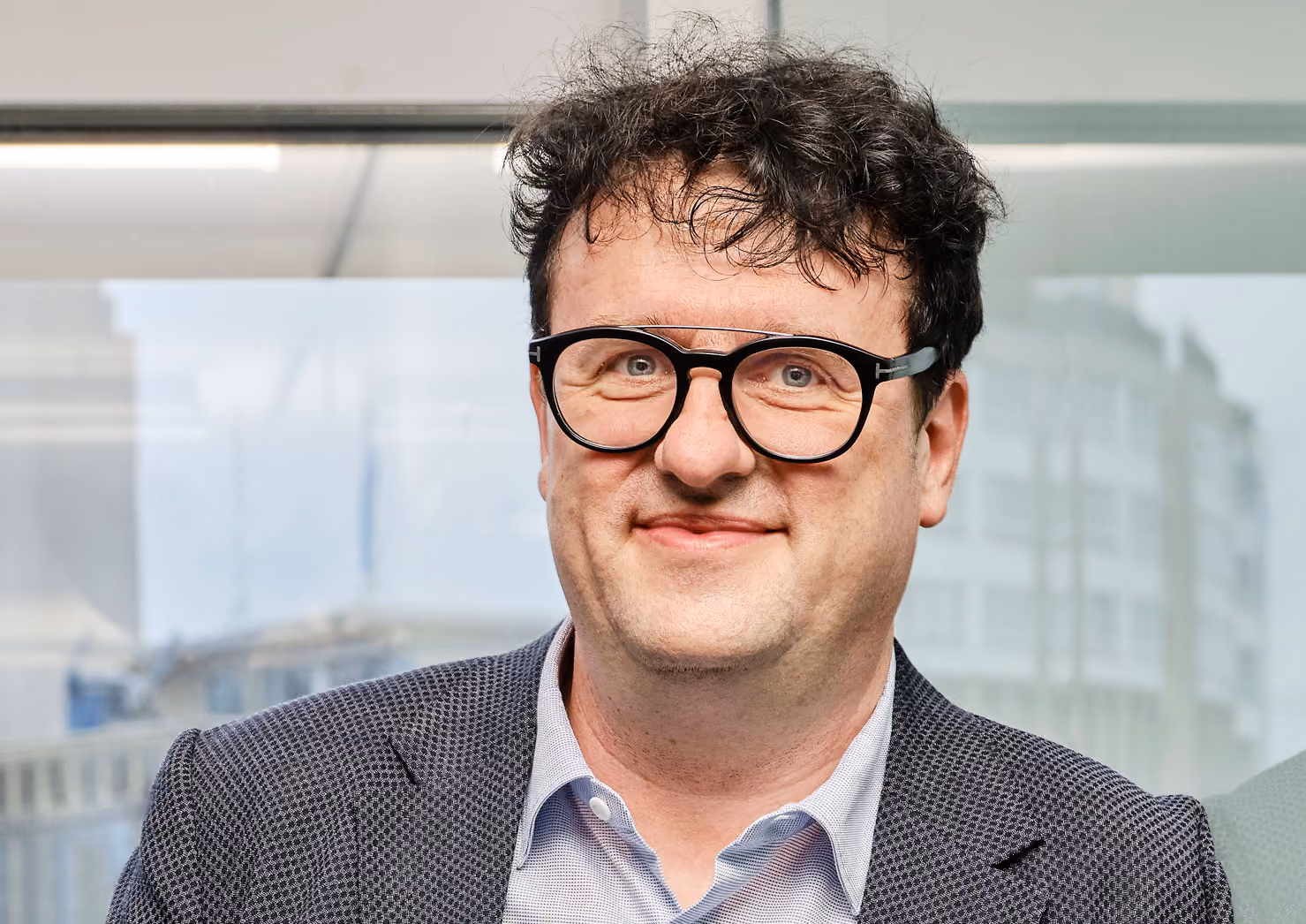 Mann mit lockigem Haar, schwarzen Brillen und blauem Sakko lächelnd vor Glasfenster.