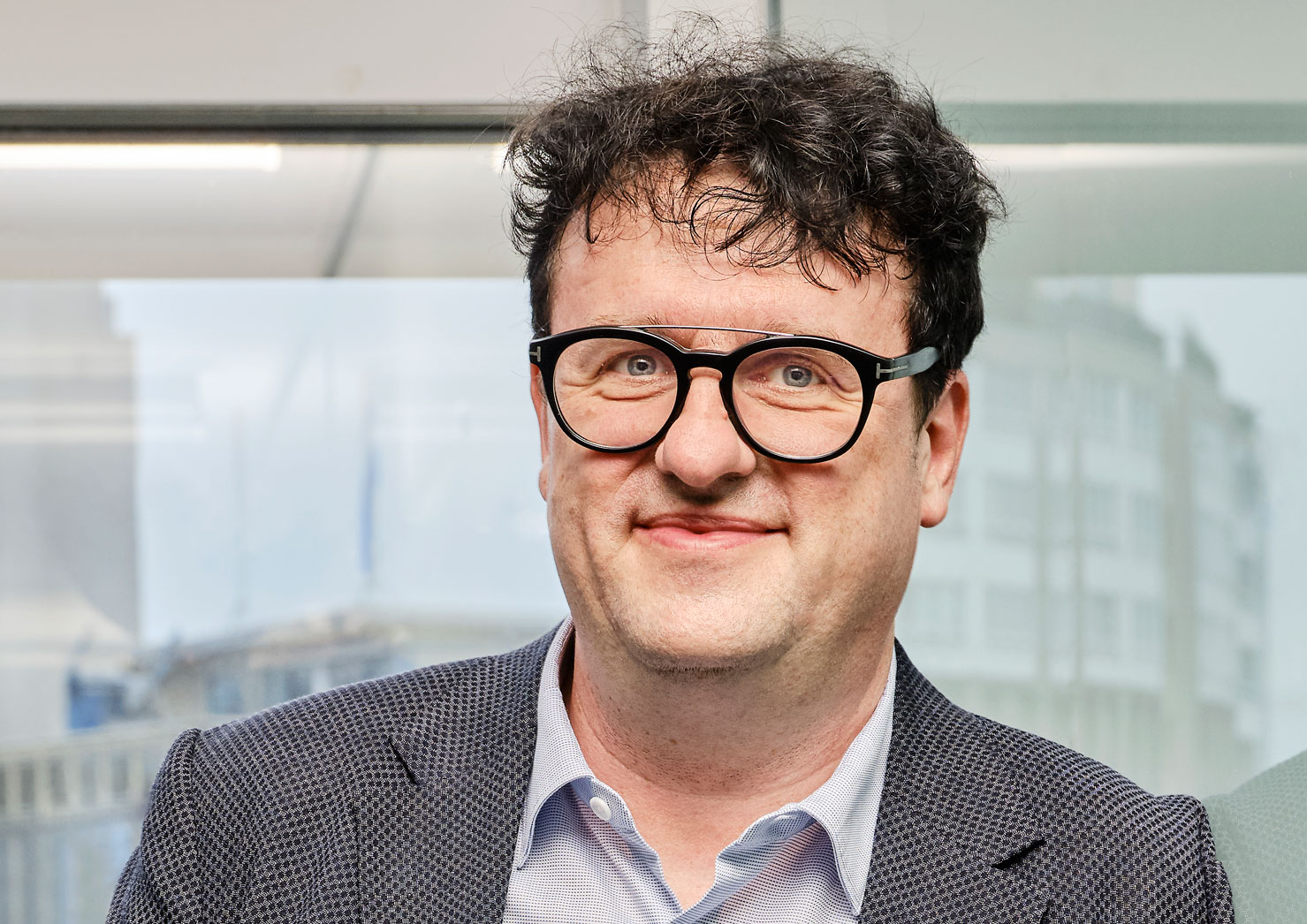 Mann mit lockigem Haar, schwarzen Brillen und blauem Sakko lächelnd vor Glasfenster.