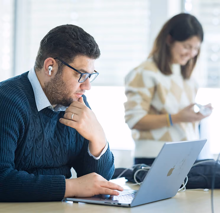 Mann mit Brille und AirPods arbeitet konzentriert an einem Laptop, Frau im Hintergrund lächelt