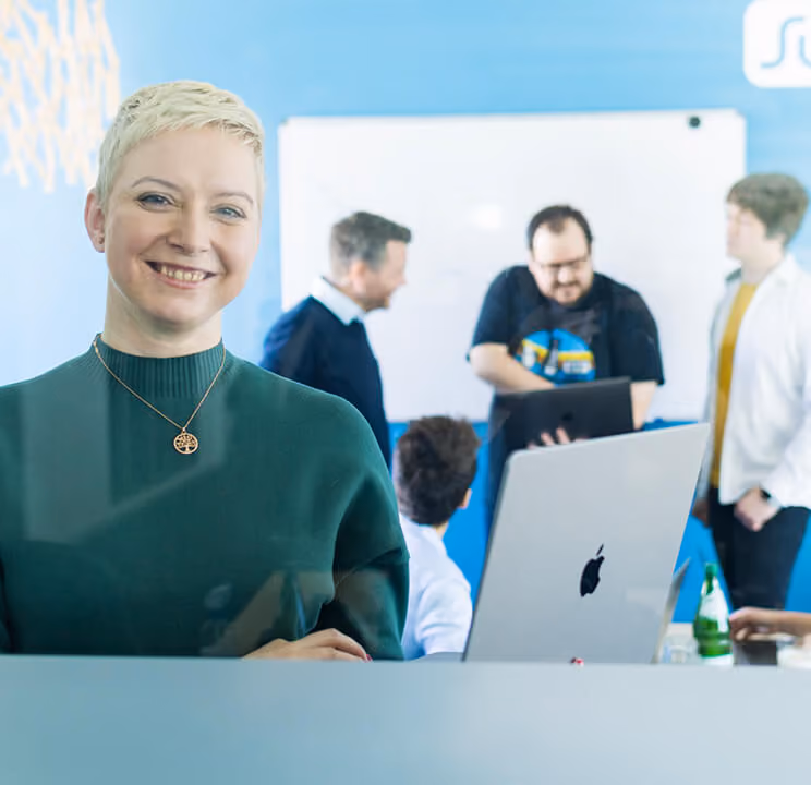 Lächelnde Frau im Vordergrund, dahinter vier Personen in einem Büro mit Laptop und Whiteboard.