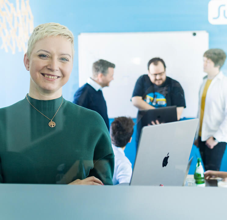 Lächelnde Frau im Vordergrund, dahinter vier Personen in einem Büro mit Laptop und Whiteboard.