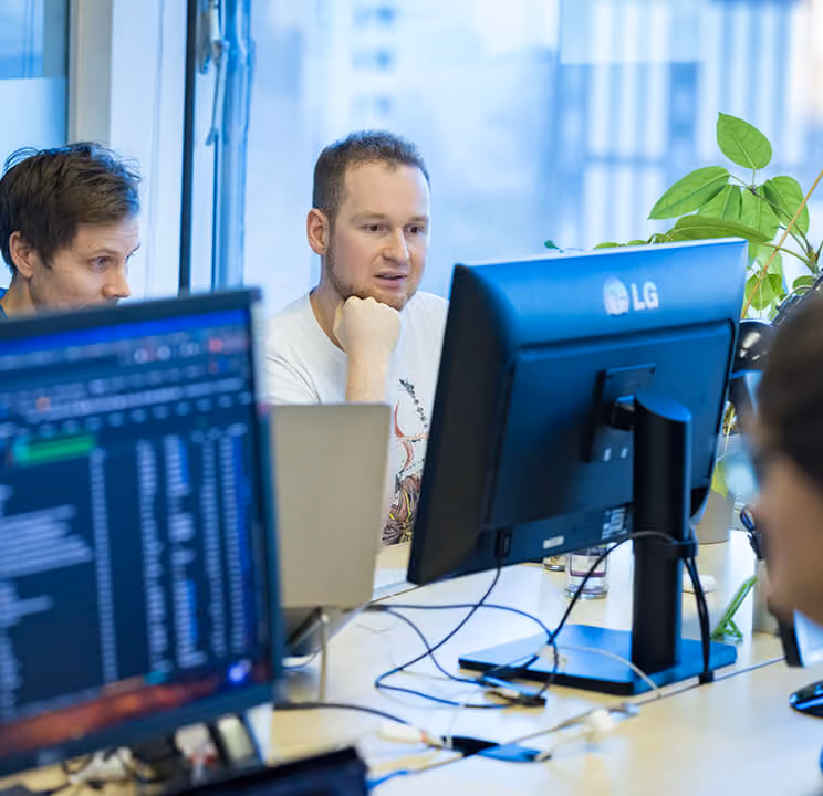 Drei Männer arbeiten zusammen an Computern in einem modernen Büro mit Pflanzen am Fenster.