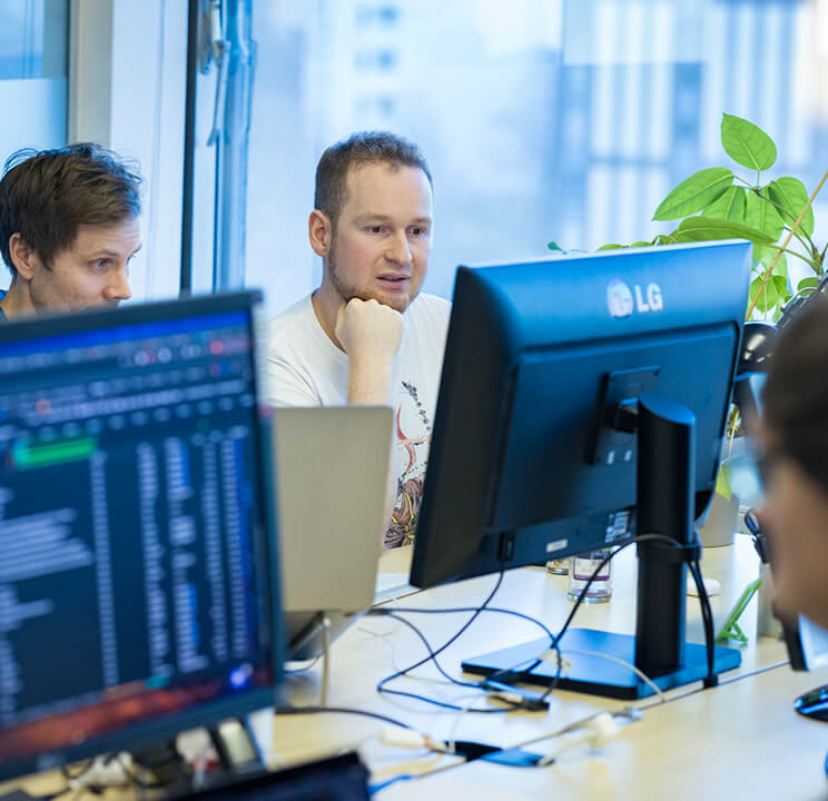 Drei Männer arbeiten zusammen an Computern in einem modernen Büro mit Pflanzen am Fenster.