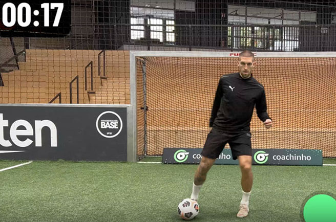 Mann im schwarzen Sportoutfit dribbelt einen Fußball vor einem Tor in der Halle.