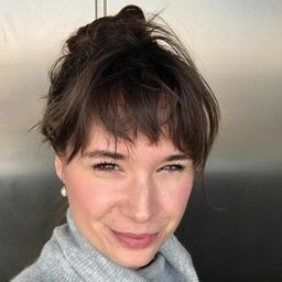 Porträt einer lächelnden Frau mit braunen Haaren und grauem Pullover vor neutralem Hintergrund.