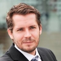 Porträt eines lächelnden Mannes mit Anzug und Krawatte vor unscharfem Hintergrund.